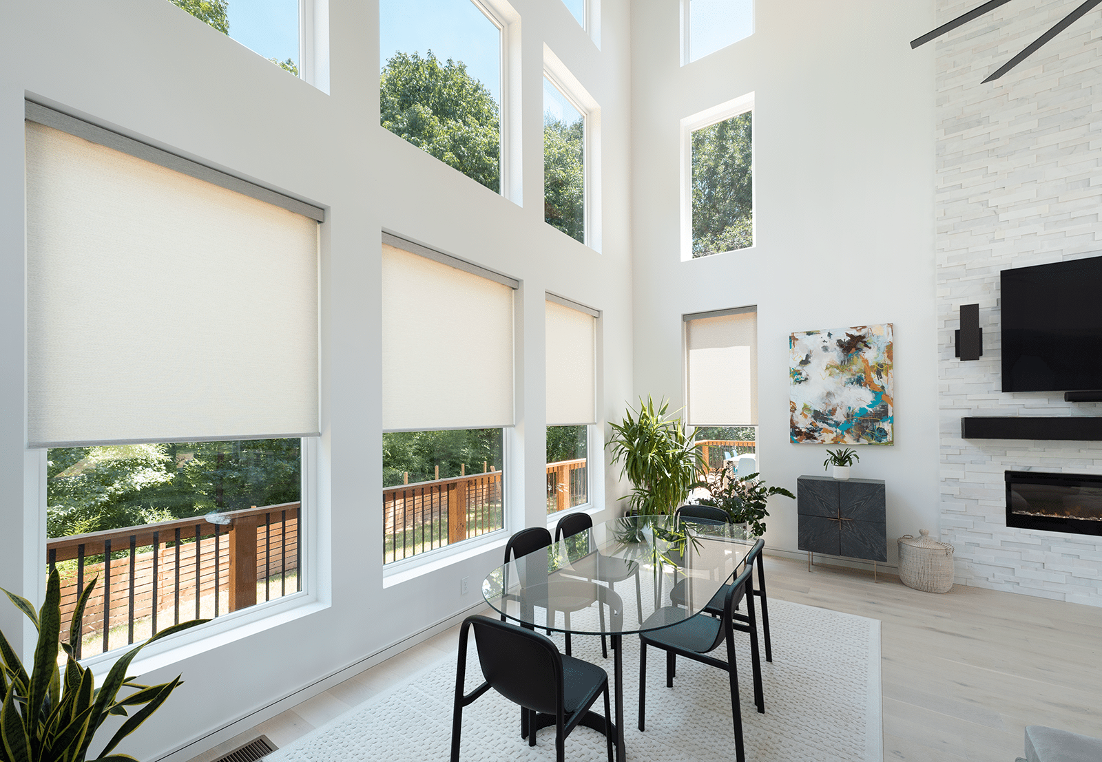 A bright and modern room featuring multiple large windows adorned with Hunter Douglas roller shades. The shades are uniformly drawn halfway, providing a balance of privacy and natural light. Outside, greenery and a wooden deck are visible.