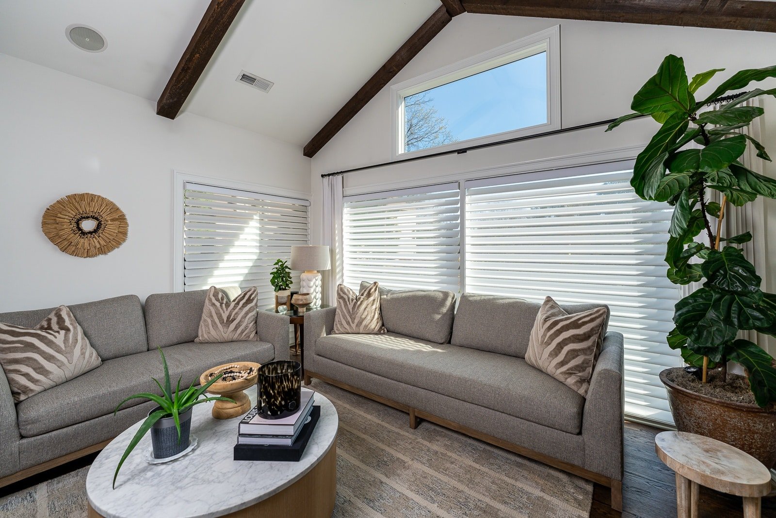 A bright living room with a gray sofa, wooden beams, sunlight coming through Silhouette shades