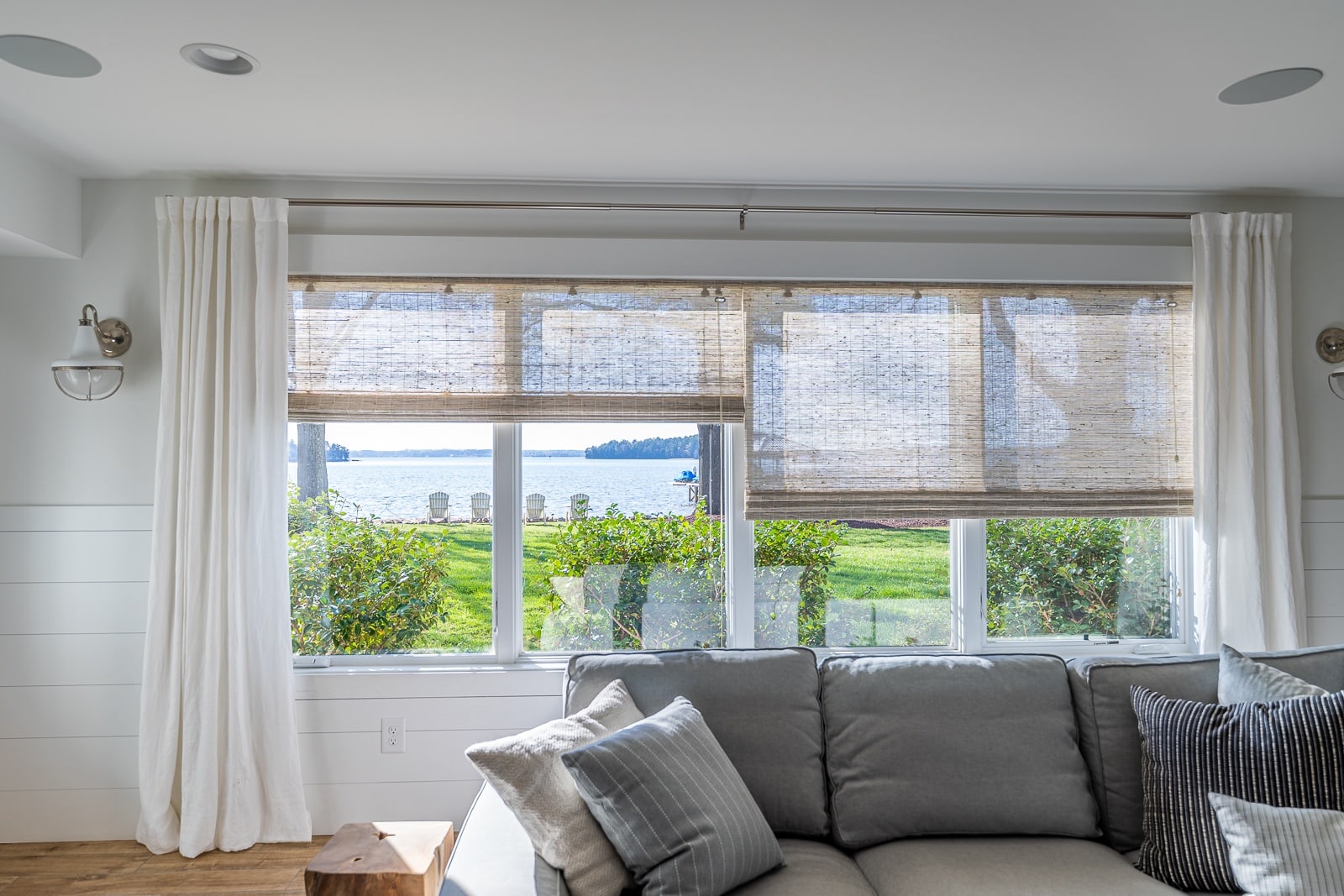 A cozy living room with a textured woven window shade, white curtains, and a serene lakeside view. The space is accented with a gray sofa and natural lighting.