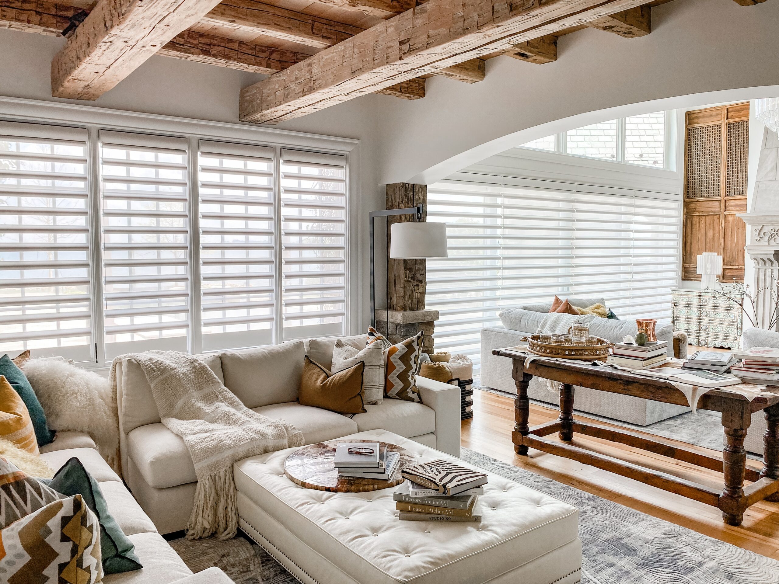 A cozy and inviting living room featuring rustic wooden beams, a plush white sofa adorned with various patterned pillows, a tufted ottoman, and a vintage wooden table. The room is illuminated by natural light filtering through the Pirouette window shadings.