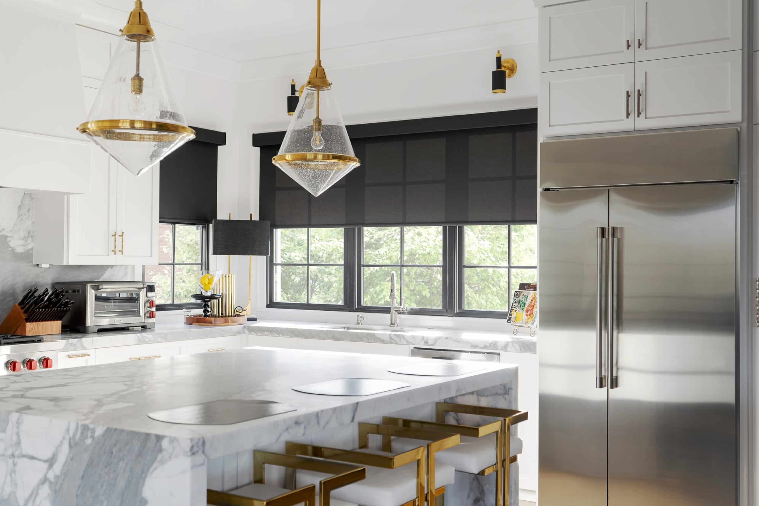 A modern kitchen with a marble island, white cabinetry, and gold accents. Black window treatments contrast the bright space, complemented by large windows showing greenery outside.