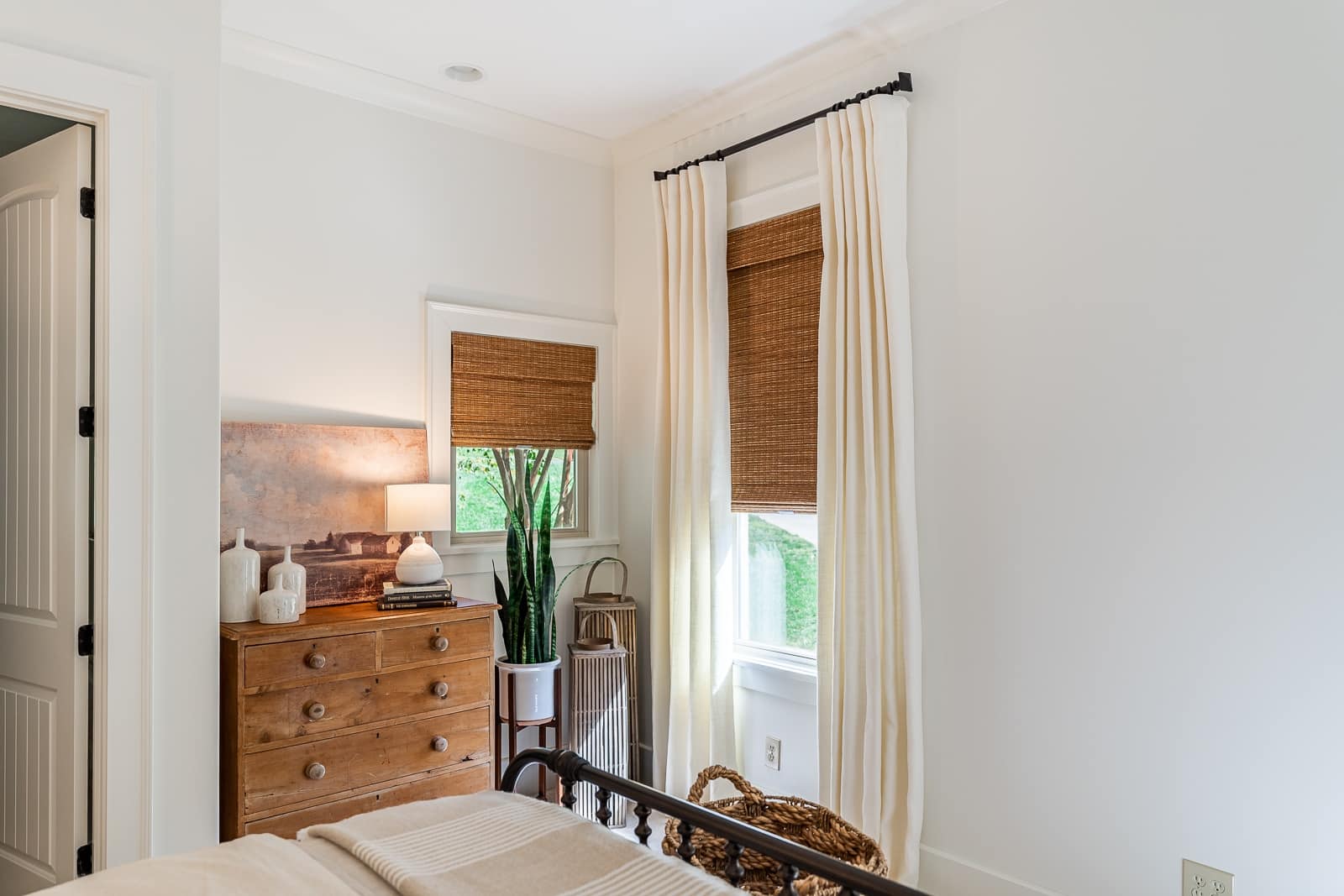 Cozy bedroom corner with wooden dresser, lamp, and woven window shades and soft white drapery