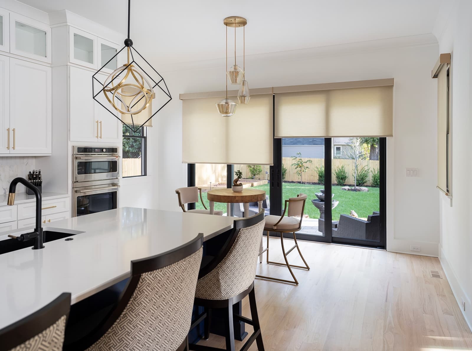 Modern kitchen and dining area showcasing a white island with patterned bar stools, geometric pendant lights, and beige roller shades on sliding glass doors that lead to a backyard