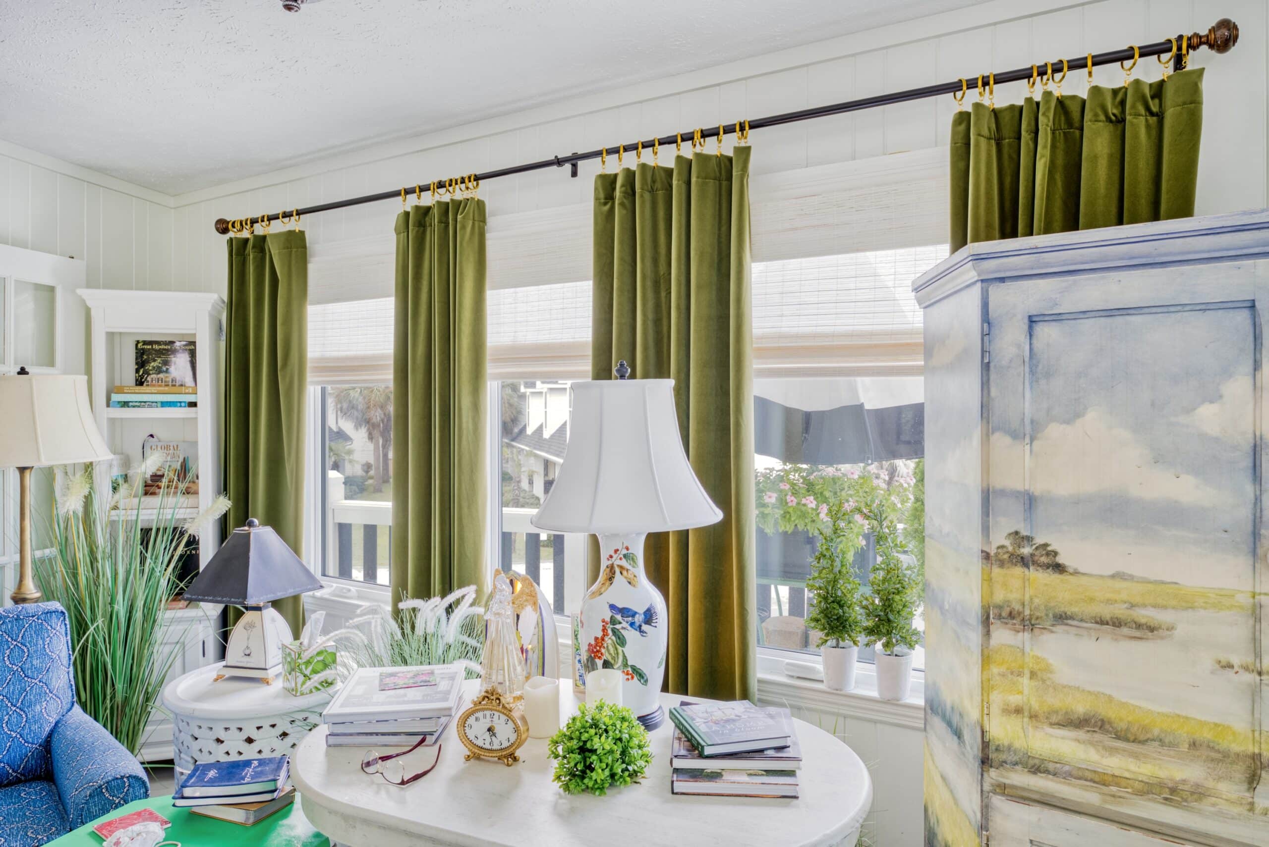 A cozy reading nook featuring green curtains overlapping white woven shades, a blue upholstered chair, and a variety of nautical and botanical decorations.