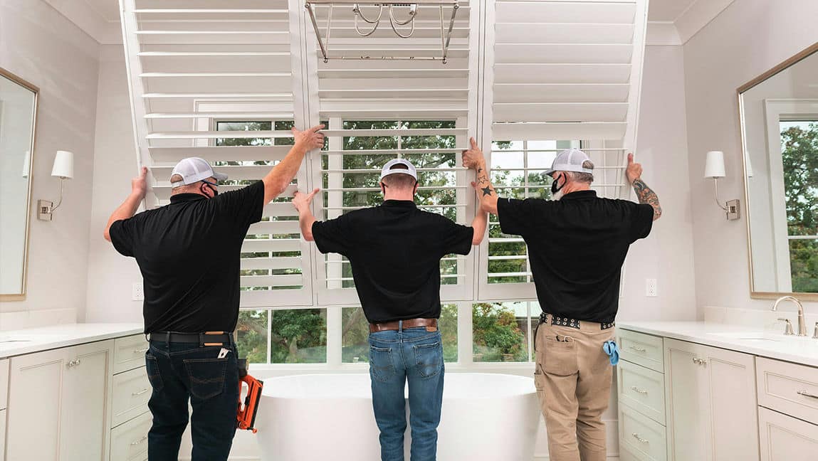 Over a bathtub, three technicians from A Shade Above maneuver a large shutter into place