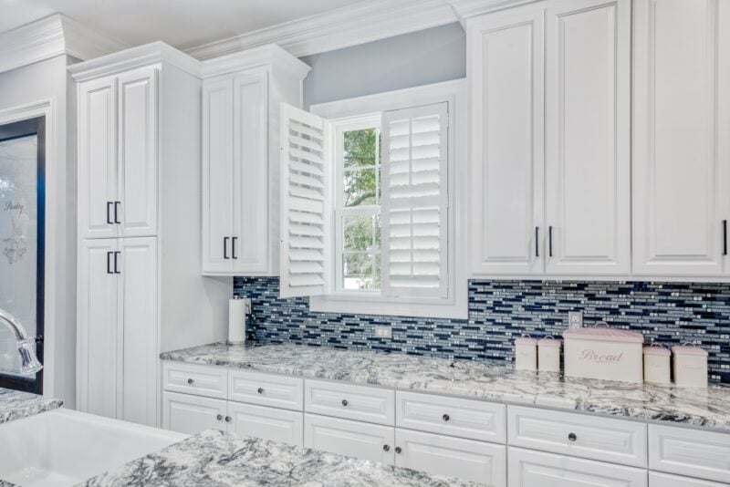 A classic kitchen with white cabinets and marble counter top with an open plantation shutter in the window 