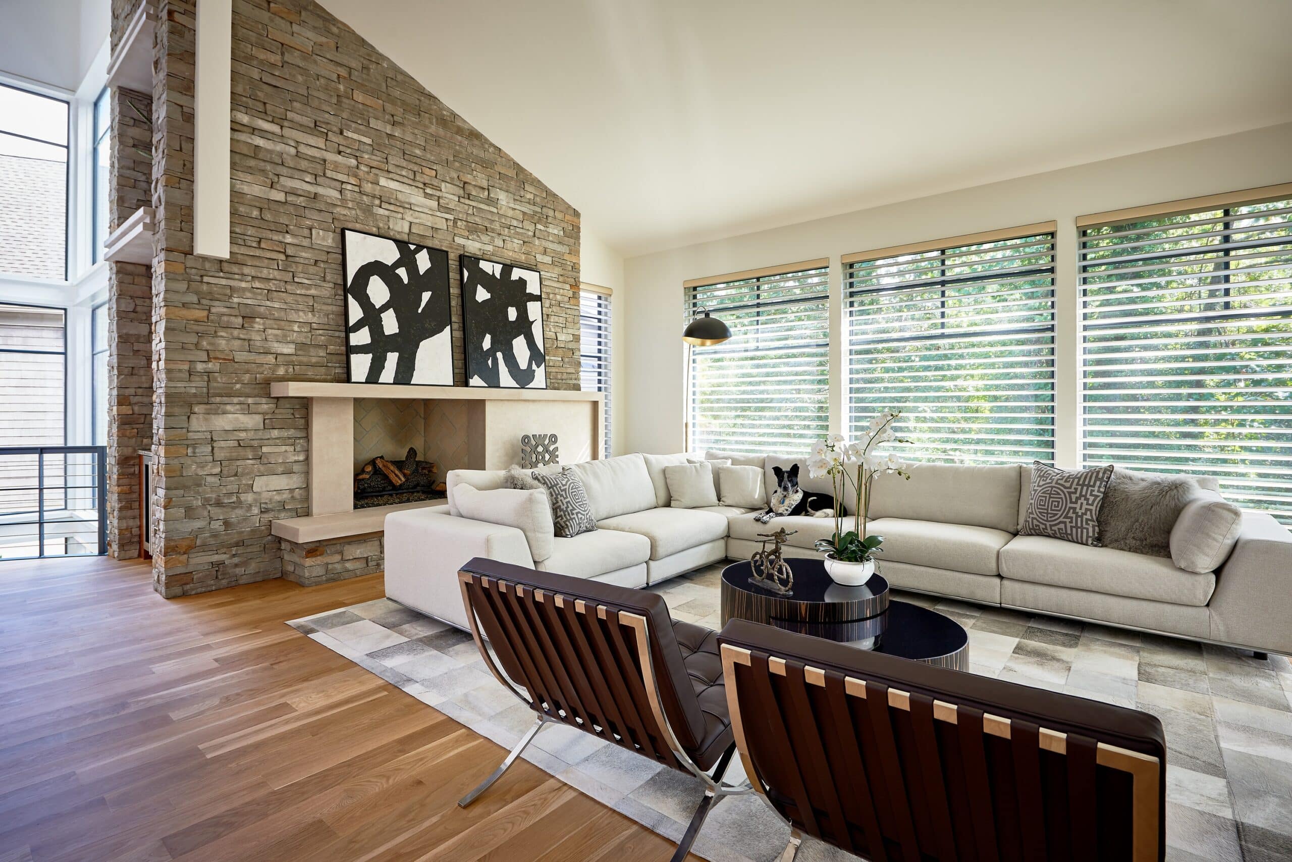 Modern living room with stone accent wall, abstract artwork, fireplace, beige sectional sofa, and large forest-view windows covered by Silhouette Shades. A dog rests on the sofa.