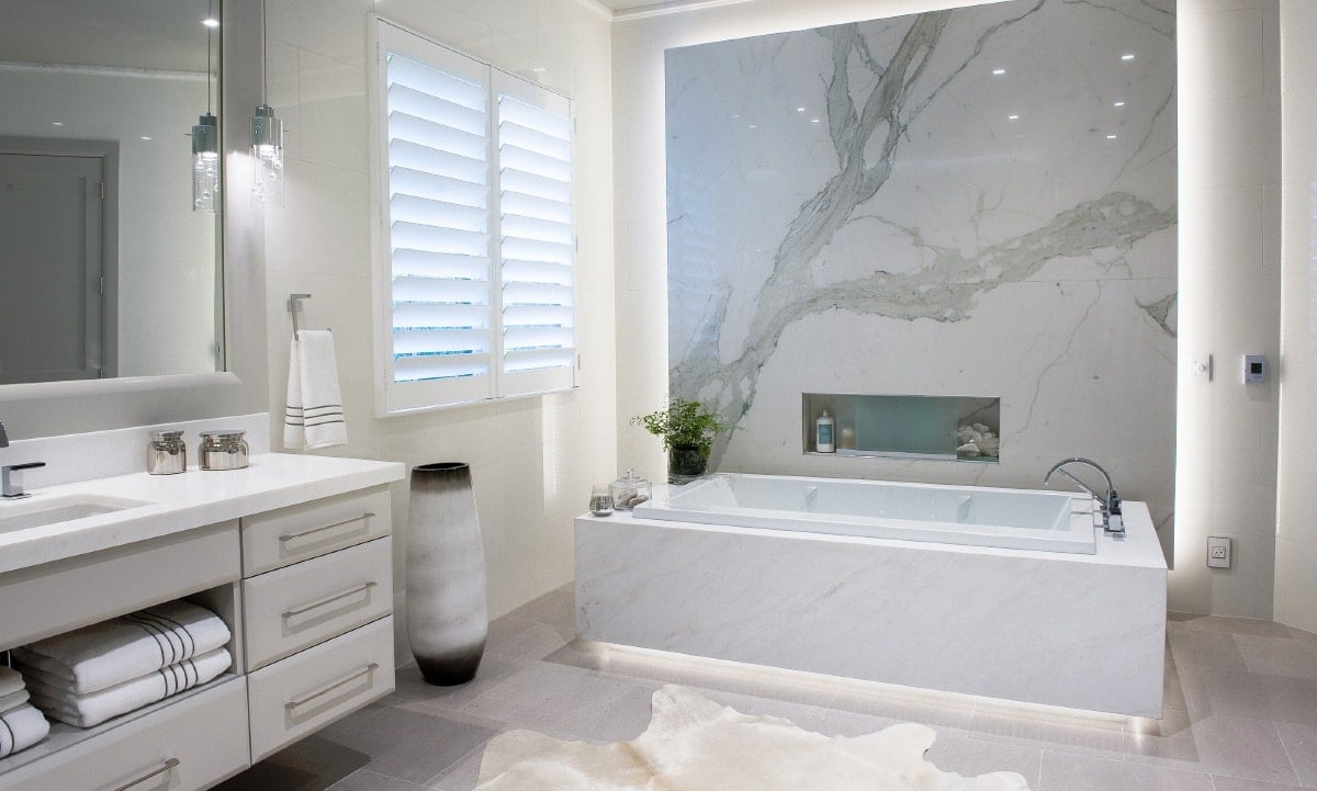 Modern bathroom with a marble wall above a tub, with a plantation shutter on the window