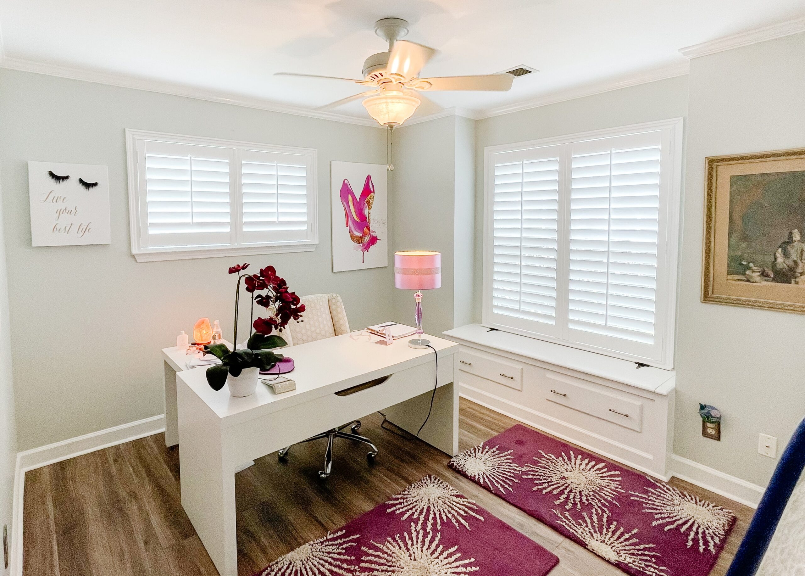 Feminine-style office space featuring a desk, accented with pink decor items, and two windows adorned with white shutters.