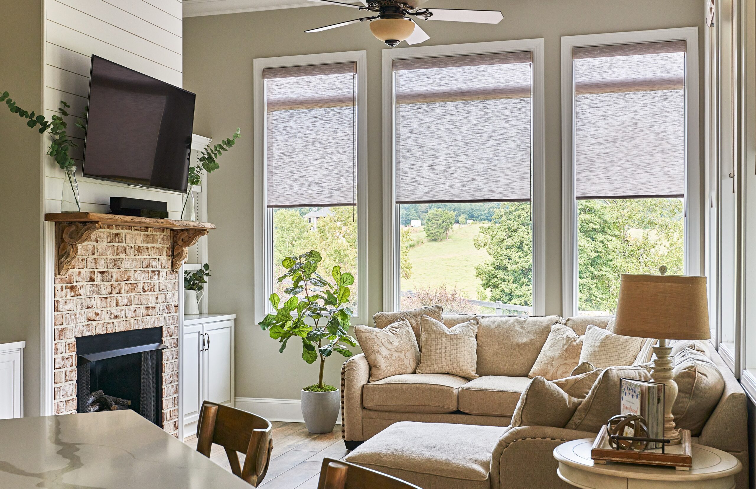 Cozy living room featuring a brick fireplace, mounted TV, neutral-toned furnishings, and large windows offering a scenic view of the outdoor country.