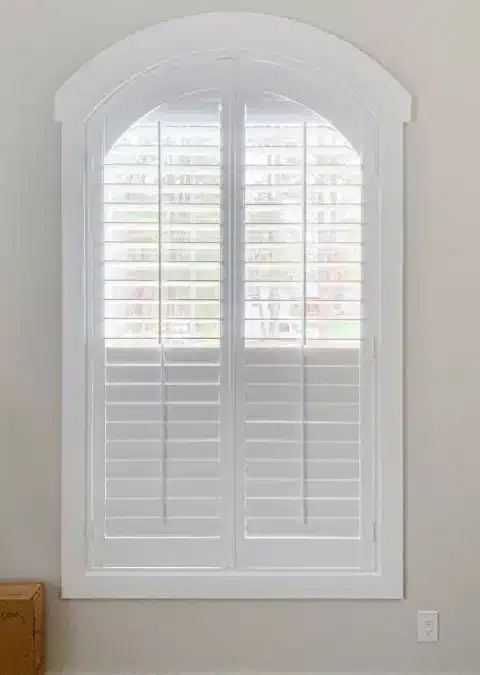 A Plantation Shutter with a curved arch top adorns a window with white trim and cream walls.