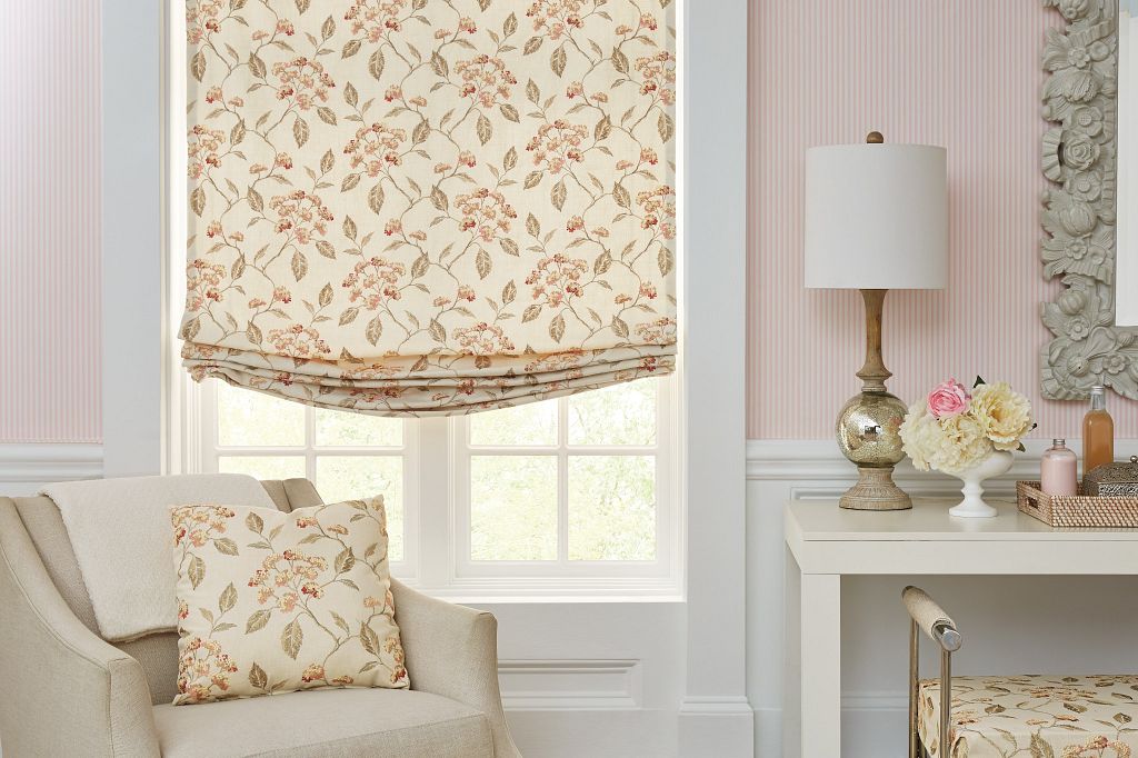 A Soft fold, relaxed roman shade in a yellow floral print hangs above a chair with a matching floral pillow in a pink bedroom. Charlotte home
