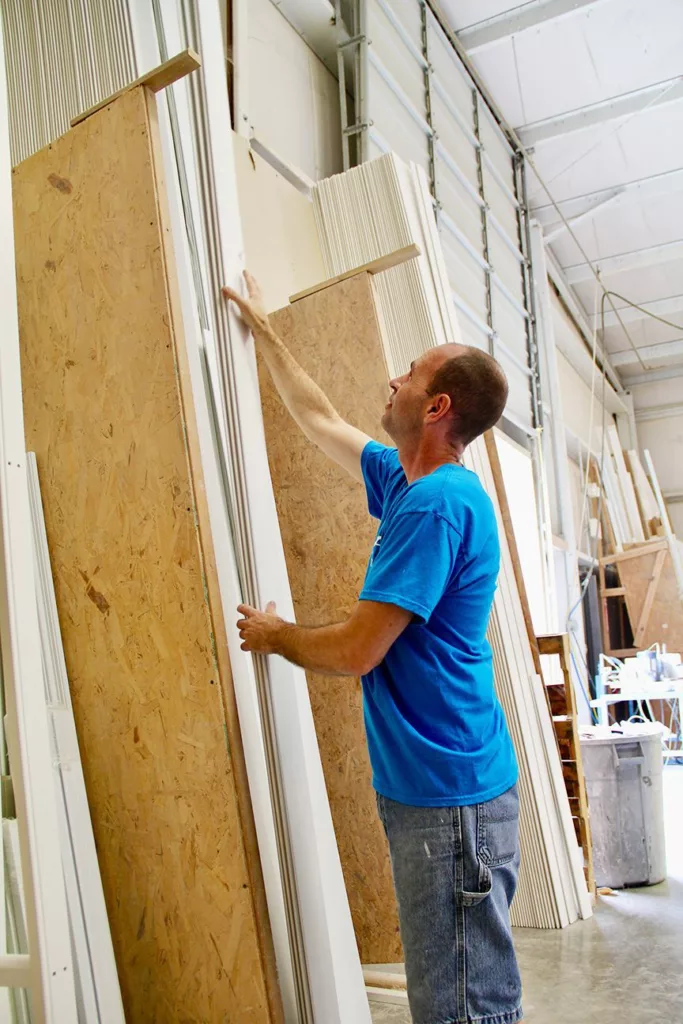 A man in a blue shirt stocks long white lumber vertically on a wall